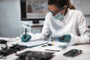 A researcher wearing a mask, safety glasses and gloves sits at a desk, looking at electronic devices and writing on a clipboard. 